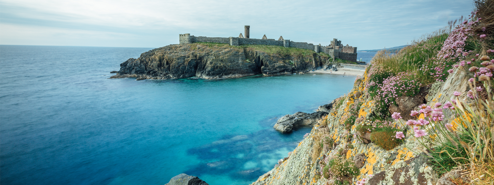 Peel Castle sitting proudly on St Patrick's Isle in Peel, Isle of Man. The image is taken from the adjacent coastline, just south of the castle. It shows the castle and St Patrick's Isle surrounded by the sparkling blue Irish Sea.